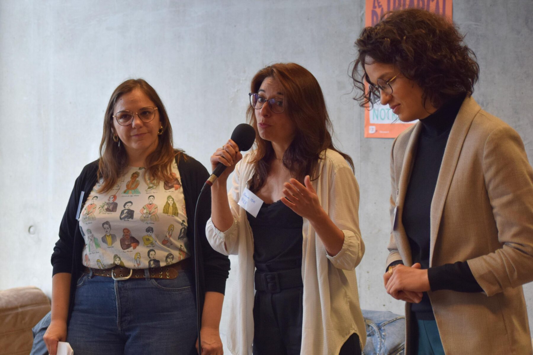 une adulte parle au micro debout sur une scène, entourée de trois autres femmes