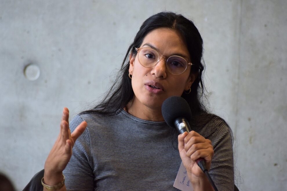 Portrait d'une femme aux cheveux longs et bruns, avec des lunettes et un haut gris, parlant au micro