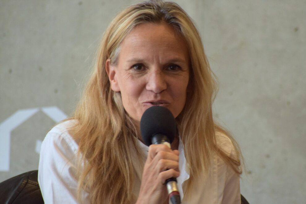 Portrait d'une femme aux cheveux longs blonds, avec un haut blanc, parlant au micro