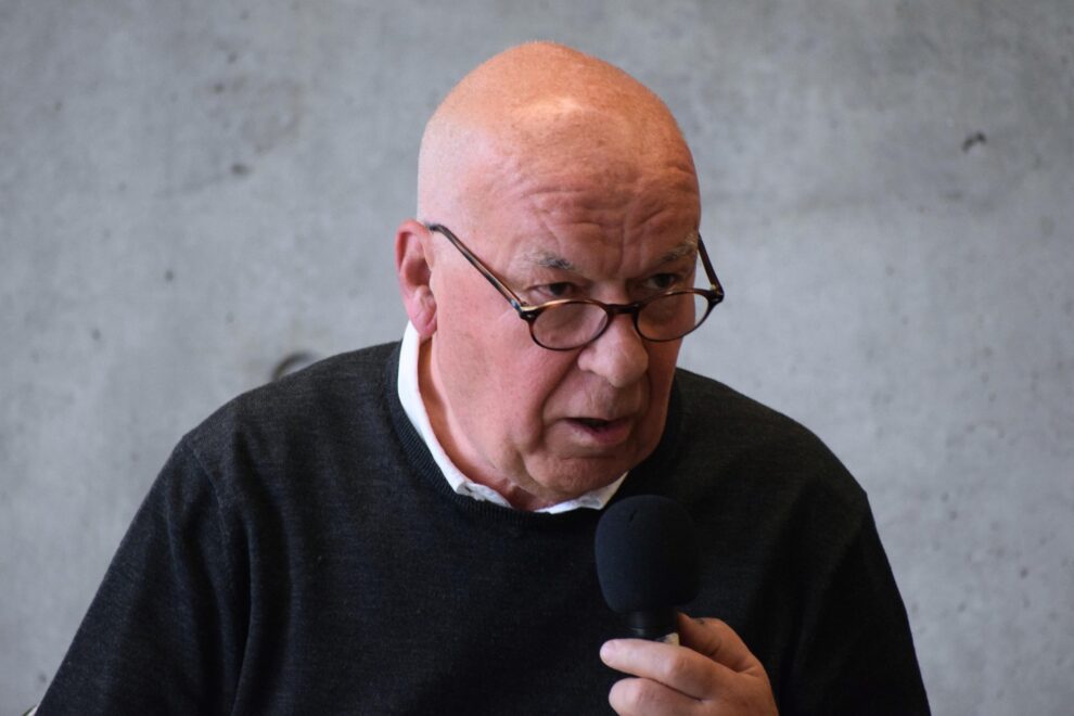 Portrait d'un homme chauve avec des lunettes et un pull noirs parlant au micro