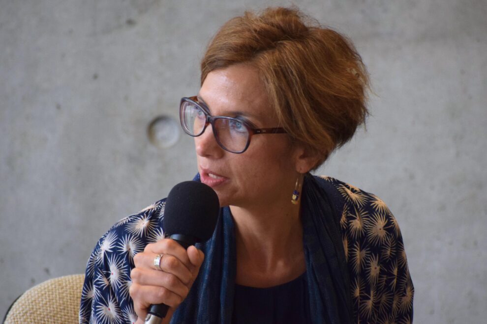 Portrait d'une femme aux cheveux châtains court, avec des lunettes et un haut bleu, parle au micro