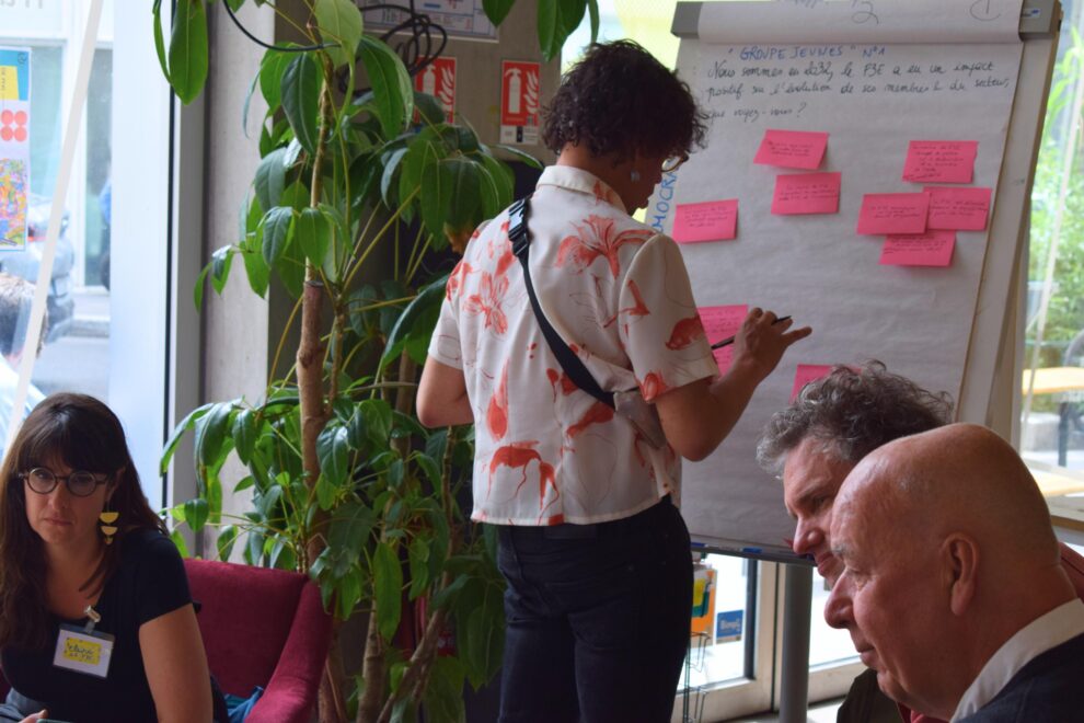 Un adulte est debout face à un tableau blanc avec des post-it roses, entouré d'autres adultes assis qui parlent entre eux
