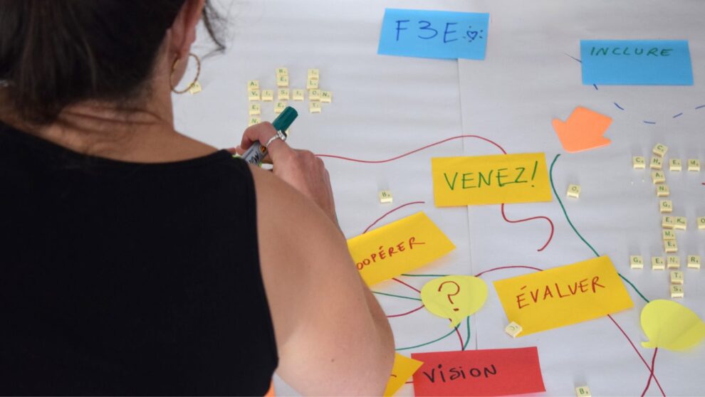 Une adulte écrit sur une large feuille blanche où se trouve des post-its avec des mots clefs tels que vision, évaluation, changement