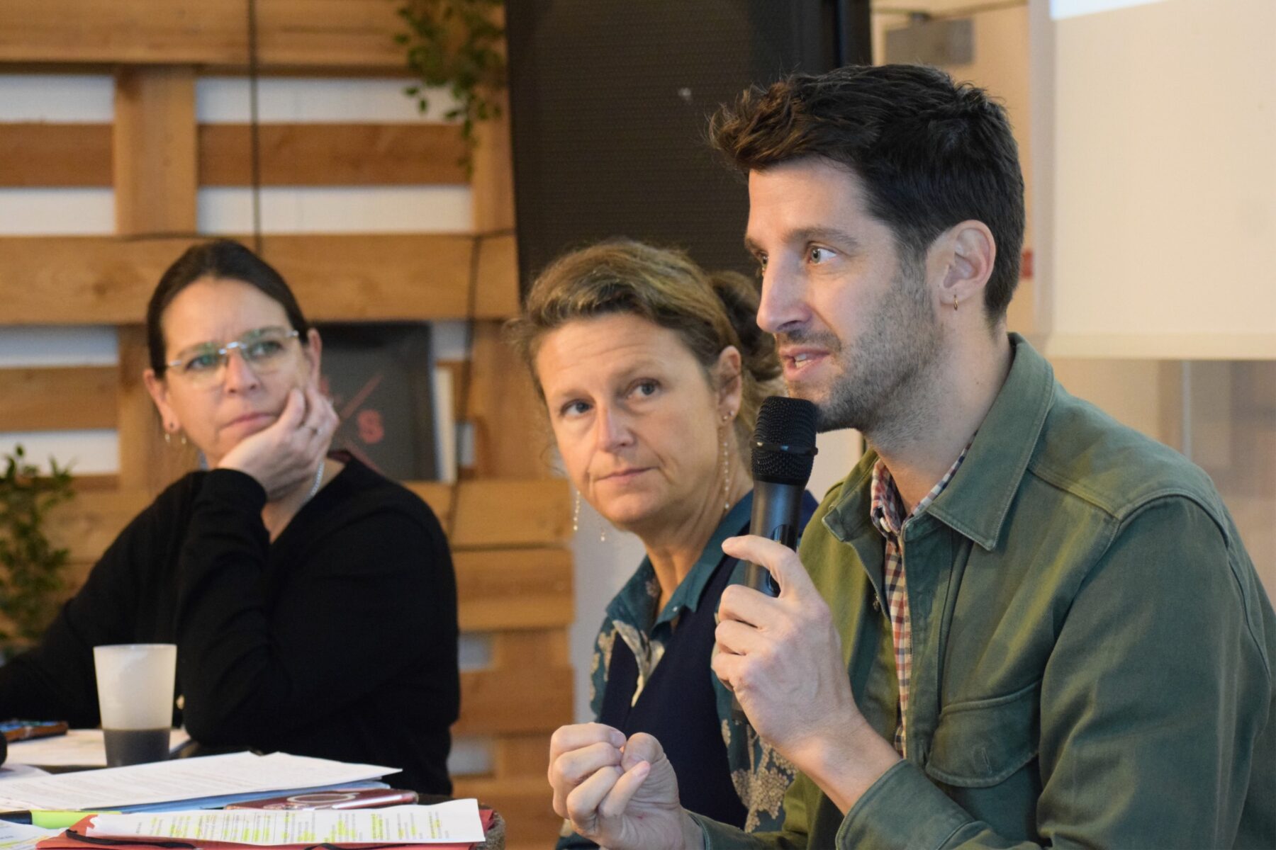 Au premier plan un adulte parle dans un micro, derrière lui deux adultes l'écoutent et le regardent