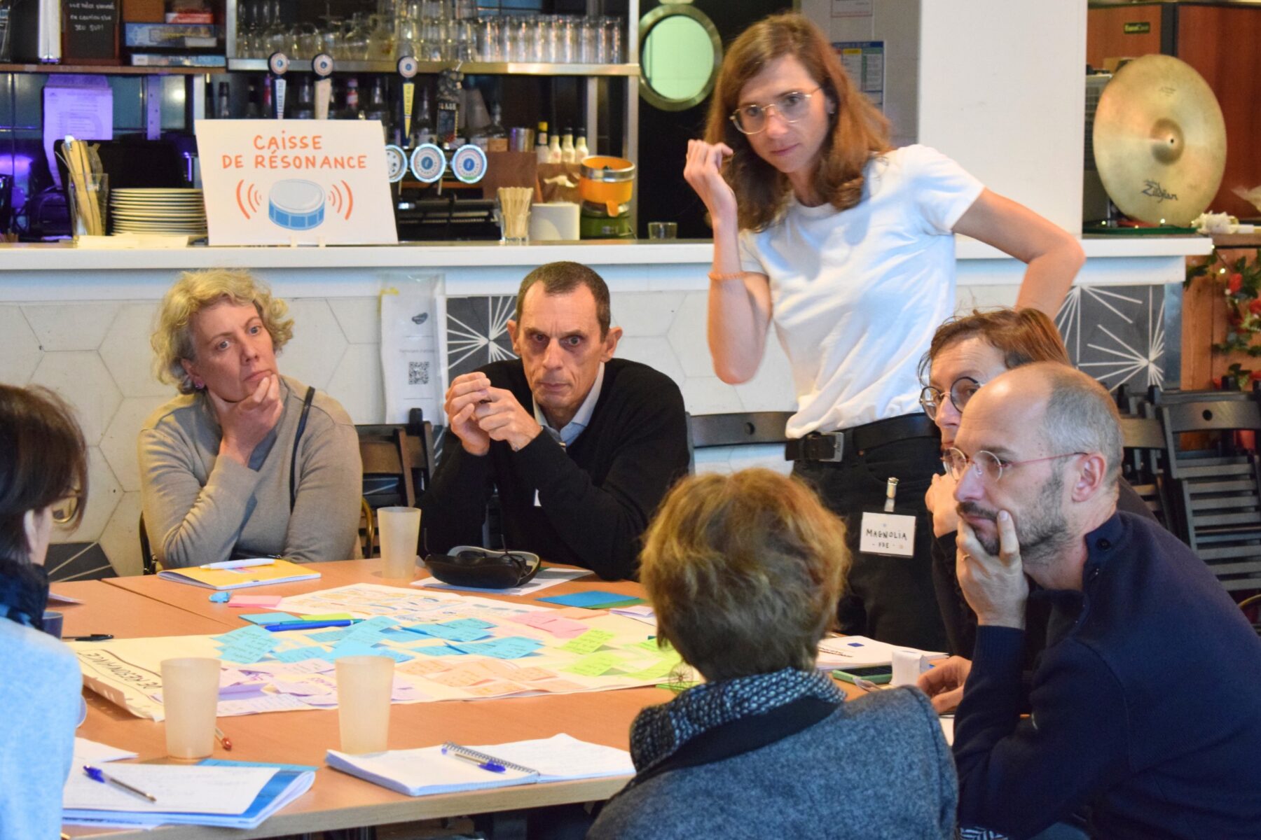Un groupe d'adultes assis autour d'une table de travail, parle et échange entre elles et eux, une adulte est debout et mène les discussions
