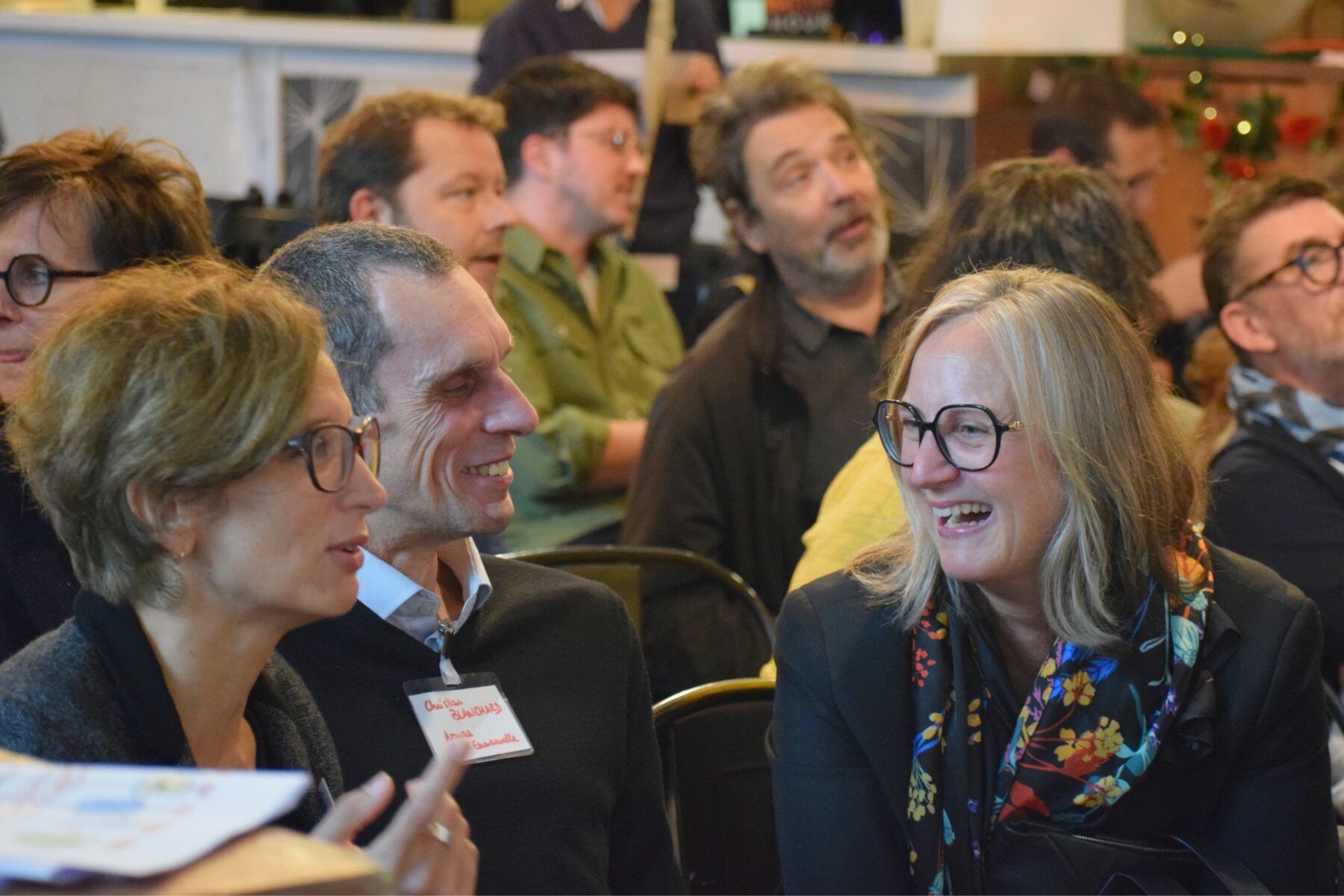 Au premier plan, trois adultes assis-e-s parlent et rient ensemble. Derrière elles et eux on voit d'autres adultes assis-e-s face à une scène