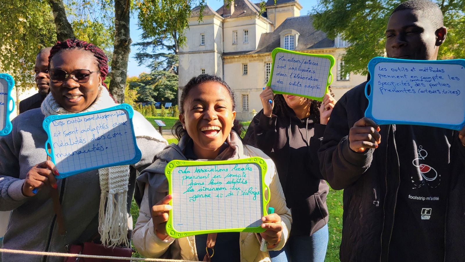 Un groupe d'adultes debout, dehors, tient des ardoises et sourient à la caméra
