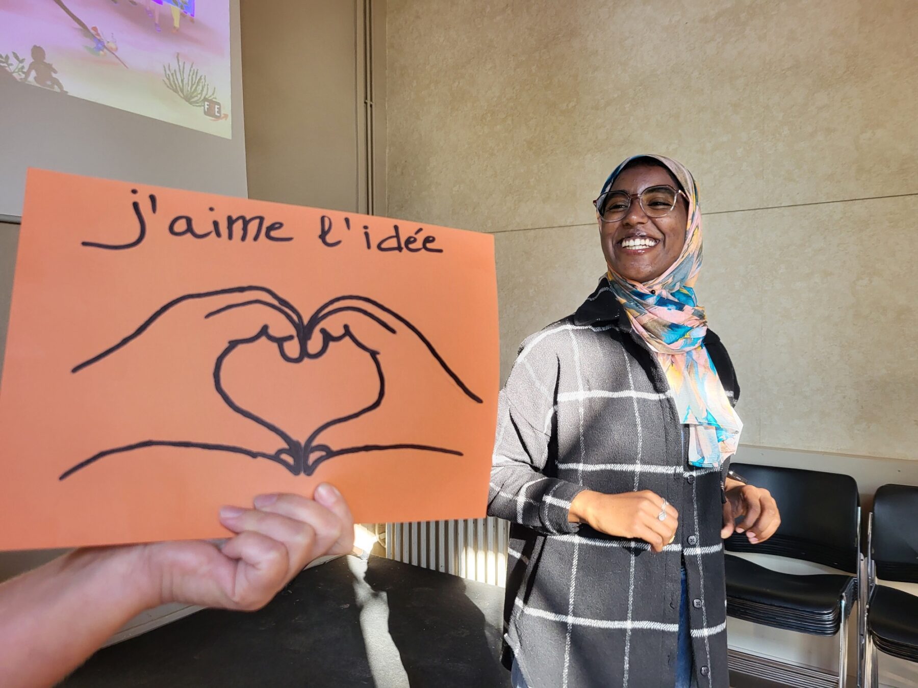 En arrière plan une adulte souris à la caméra, devant un post-it orange avec le texte "j'aime l'idée"