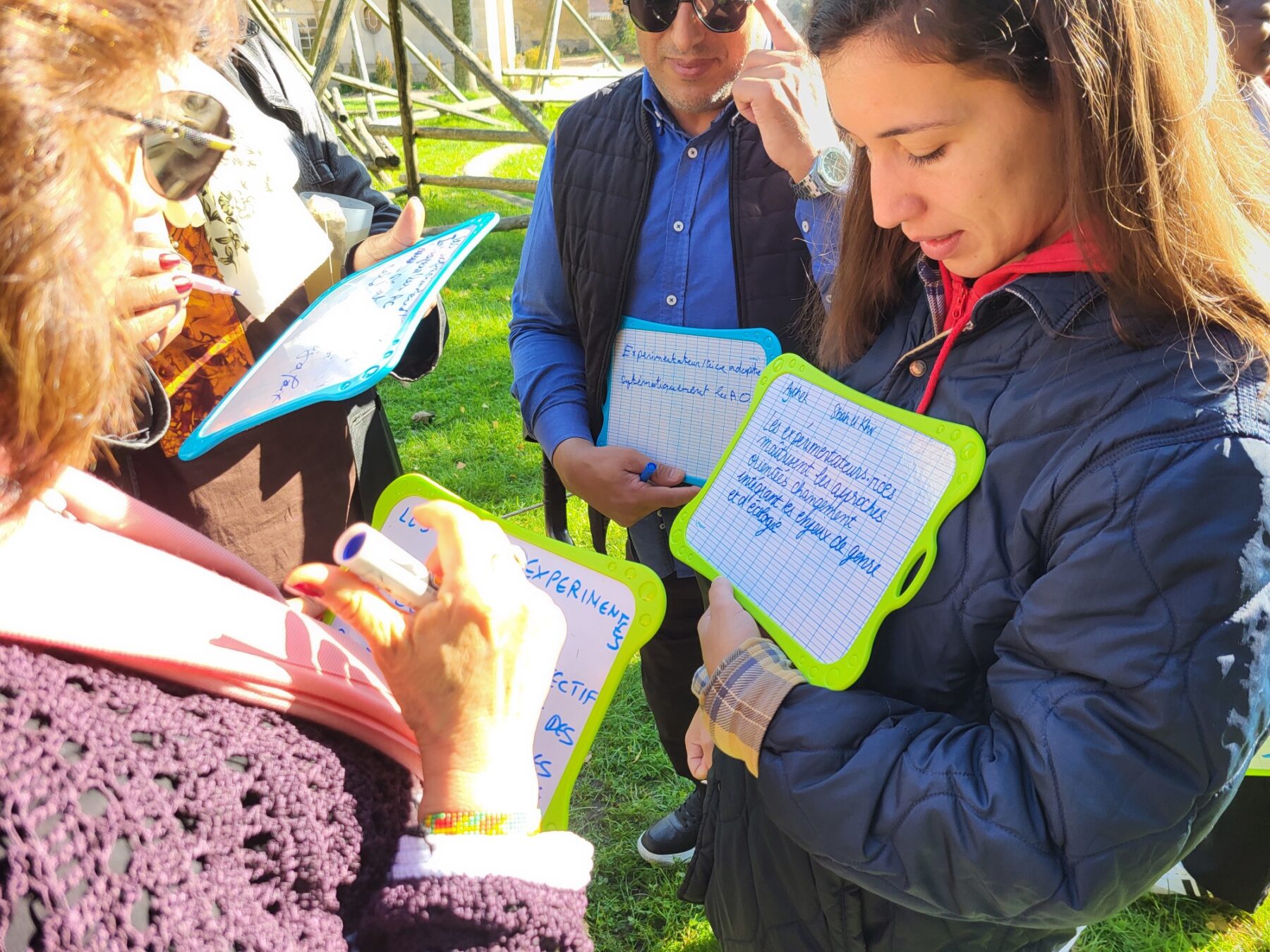 Un groupe d'adultes debout, dehors, tient des ardoises et échangent des idées