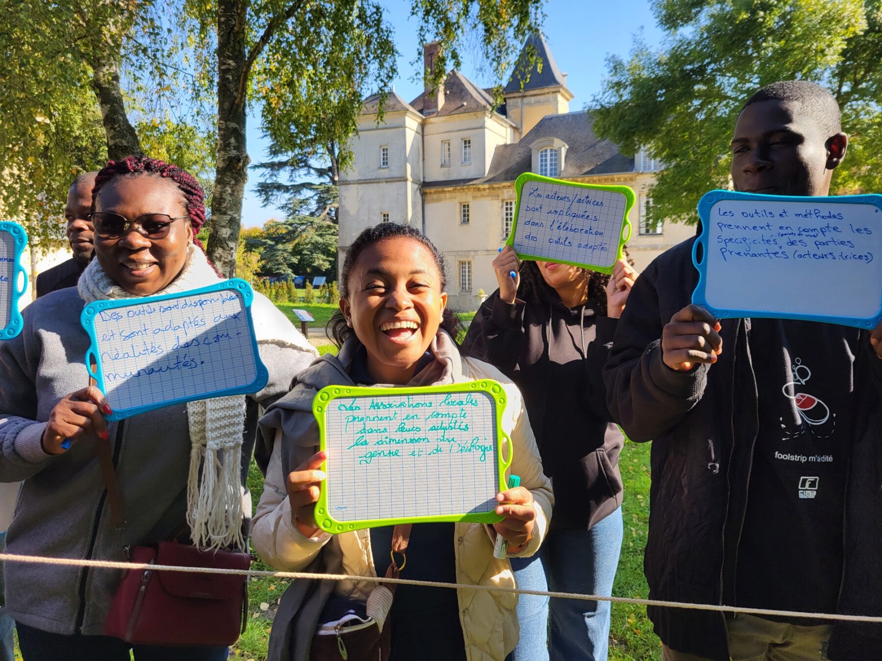 Un groupe d'adultes debout, dehors, tient des ardoises et sourient à la caméra