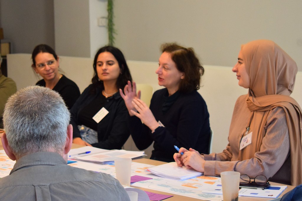 Un groupe d'adultes assis-e-s autour d'une table, écoute une adulte parler